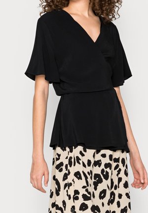 Vrouw draagt een zwarte V-halsblouse met vliegende mouwen en een beige rok met zwarte dierenprint en knopen aan de voorkant.