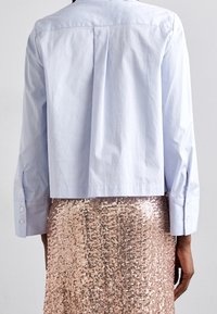 Avkortad ljusblå skjorta med långa ärmar, knappmanschetter och ryggveck, i kombination med en glittrande rosa paljettkjol.