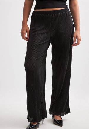 Kobieta ubrana w czarne spodnie o wysokim stanie, plisowane i szerokie nogawki, zestawione z czarnymi szpilkami z migdałowym noskiem, stojąca na jednolitym tle.