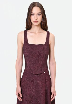 Femme aux longs cheveux bruns portant une robe sans manches texturée de couleur violet foncé avec un col carré, debout devant un fond blanc uni.