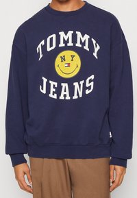 Námořnická modrá mikina vyrobená z bavlny, s bílým nápisem "TOMMY JEANS" a grafickým návrhem žlutého smajlíka. Hladké manžety a lem.