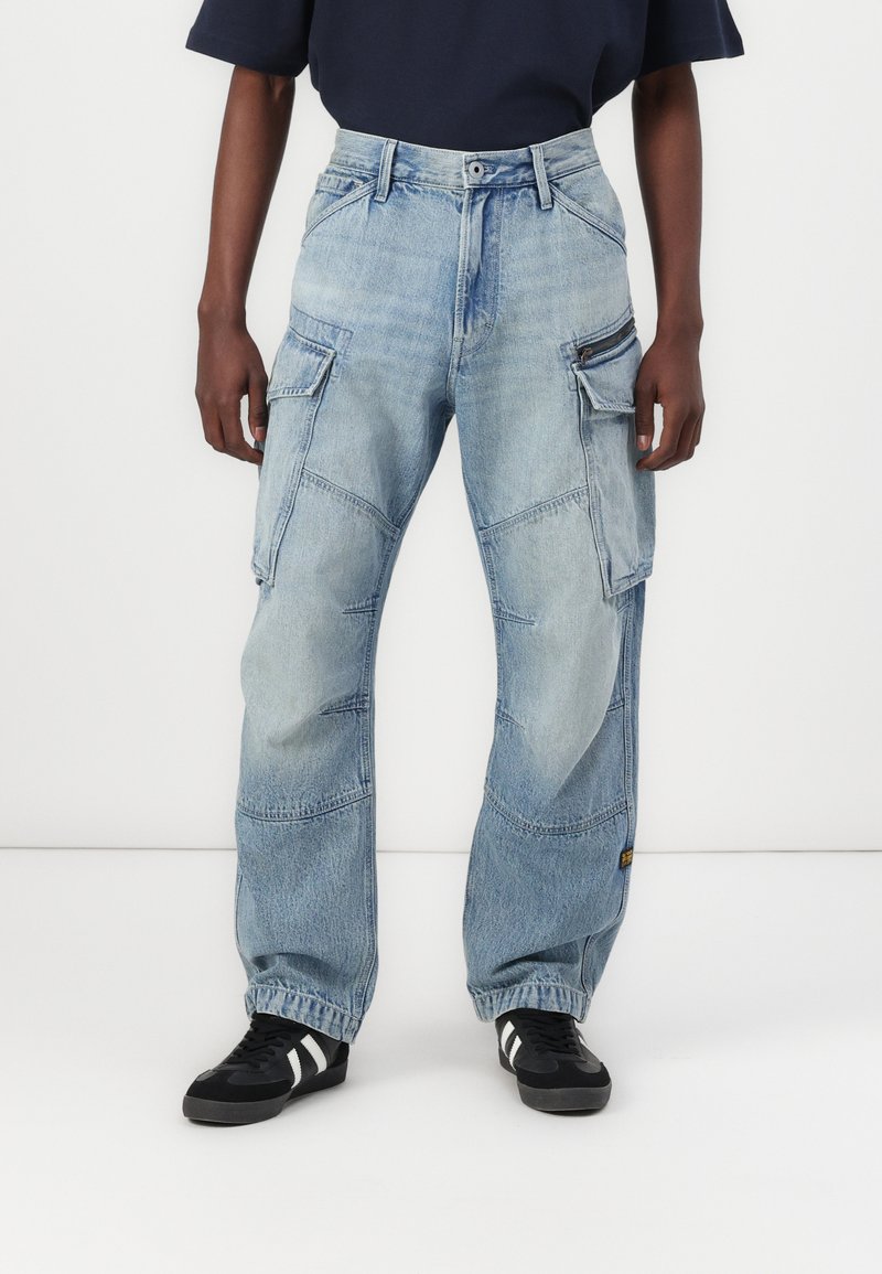 Γαλάζια τζιν cargo παντελόνια με ελευθερία στη γραμμή, πολλαπλές τσέπες, ξεθωριασμένη υφή και αντίθεση στη ραφή. Φορεμένα με μαύρα αθλητικά παπούτσια.