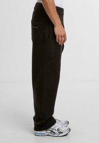 Pantalones de forro polar negros con una textura suave, que cuentan con un bolsillo lateral y una cinturilla elástica. Se llevan con zapatillas deportivas grises y blancas.