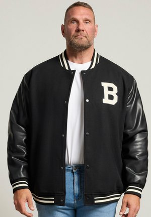 Homme portant une veste universitaire noire avec des manches en cuir et une grande lettre blanche B sur la poitrine, par-dessus une chemise blanche et un jean bleu.
