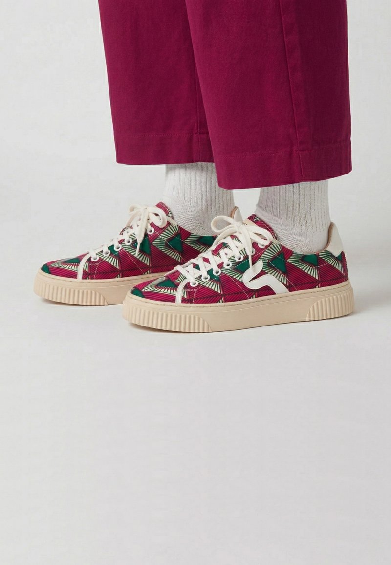Des pieds portant des baskets à motifs avec un motif géométrique rouge, vert et blanc, des chaussettes blanches et un pantalon rouge court sur un fond uni.