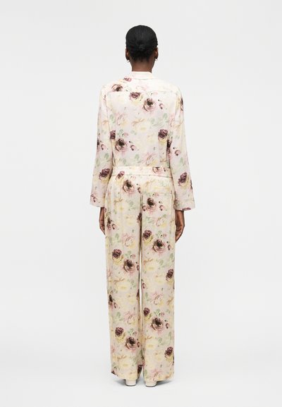 Γυναίκα με σκουρόχρωμο δέρμα και μαλλιά δεμένα πίσω, φορώντας εκρού μακρυμάνικη μπλούζα με floral μοτίβο και φαρδύ παντελόνι, στέκεται με την πλάτη γυρισμένη σε λευκό φόντο.