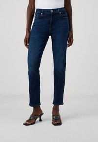 Jean en denim bleu foncé avec une coupe ajustée, style cinq poches standard et légèrement extensible. Présente des boutons en laiton et un ourlet court.
