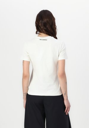 Femme aux longs cheveux bruns portant un T-shirt blanc à manches courtes avec "KARL LAGERFELD" sur le haut du dos et un pantalon noir, debout sur fond blanc.