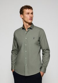 Camicetta verde oliva a maniche lunghe con colletto, bottoni frontali, vestibilità slim e un piccolo logo blu scuro sul lato sinistro del petto. Tessuto dalla superficie liscia.