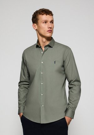 Chemise à manches longues vert olive avec un col, des boutons sur le devant, une coupe ajustée et un petit logo bleu foncé sur la poitrine gauche. Texture de tissu lisse.