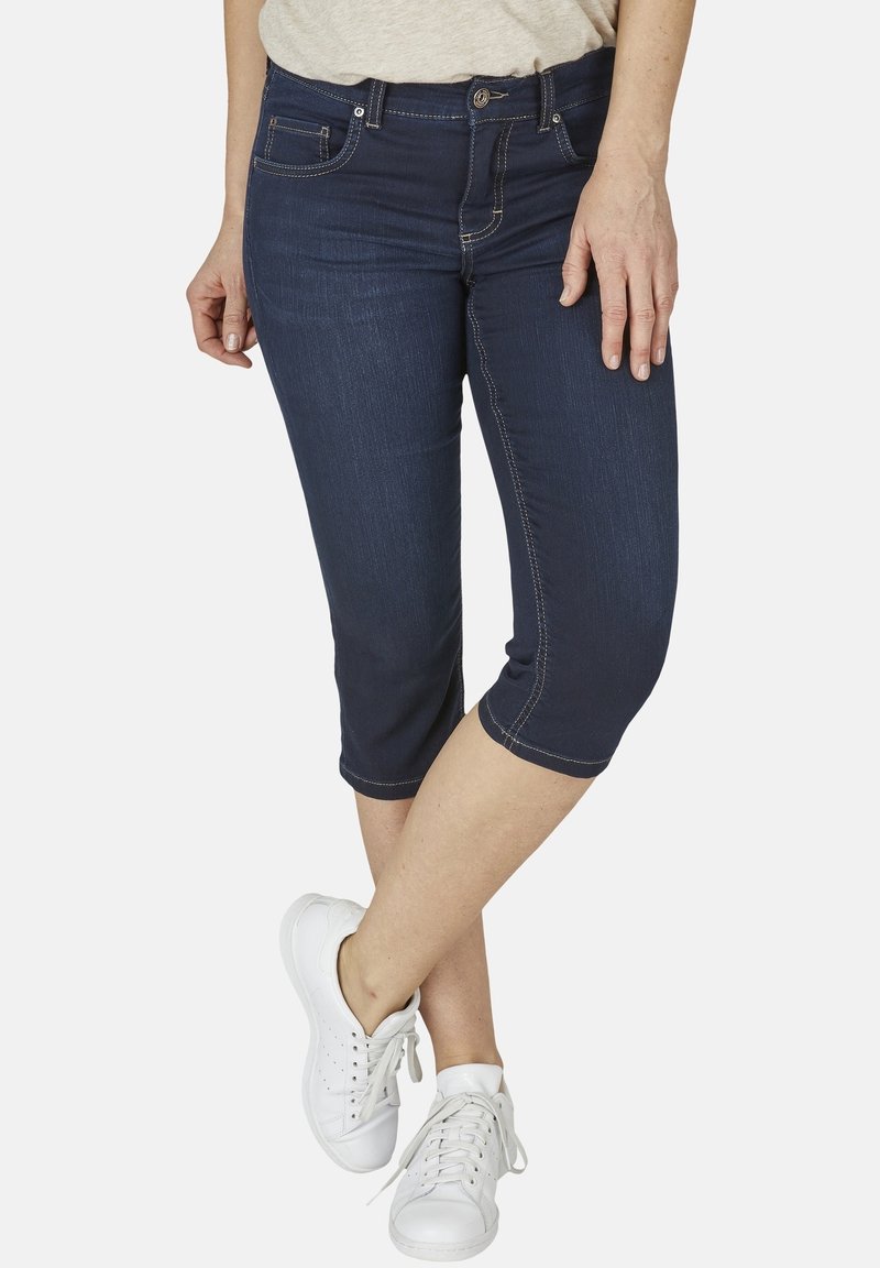 Délkové džínové capri kalhoty tmavě modré barvy s přiléhavým střihem, kontrastním prošitím a kapsami, spárované s bílými teniskami.