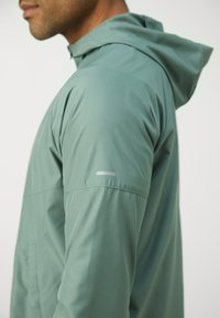 Veste légère avec capuche de couleur vert muted ; présente une texture lisse et soyeuse ainsi qu'une fine bande réfléchissante sur la manche gauche.