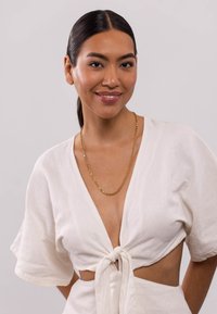 Weißes Leinenoberteil mit kurzen Ärmeln und geknoteter Vorderseite. Goldene Kettenhalskette. Heller Hintergrund, glatte Textur und einfaches Design.