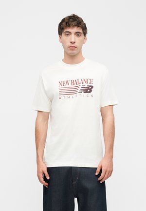 Giovane uomo rivolto in avanti che indossa una maglietta bianca New Balance Athletics e jeans blu scuro su uno sfondo bianco semplice.
