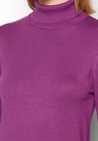 Gros plan d'une personne portant un pull à col roulé côtelé violet, montrant l'encolure et le haut du torse.