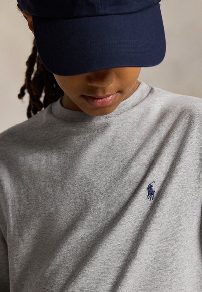 Oseba, ki nosi mornarsko kapo in svetlo sivo majico z majhno temno modro vezenino igralca polo na prsih, z glavo nagnjeno navzdol.