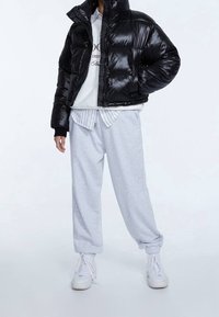 Giacca a vento nera con finitura lucida, design cropped e colletto alto; abbinata a pantaloni della tuta grigi chiari e sneakers bianche.