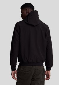 Jachetă neagră cu glugă, cu o textură netedă și manșete elastice, având un design ajustat și cusături la umeri, asociată cu pantaloni cargo verzi măslinii.