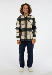 Man met krullend haar die glimlacht, gekleed in een beige en zwart geruit overhemd met knopen, donkerblauwe jeans en witte sneakers, staand op een witte achtergrond.
