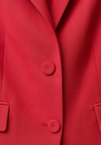 Punainen bleiseri, jossa on muotoiltu leikkaus, halkiot, kaksi etutaskua ja kaksi pyöreää nappia edessä kiinnittämiseksi. Sileä kankaan rakenne.