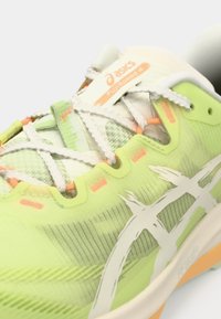Green and white athletic shoe with textured mesh upper, orange accents, and reflective details. Features lacing system and branded logo.