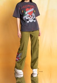 Grafické šedé tričko s designem lebky a srdce. Olivově zelené cargo kalhoty s výšivkou orla. Bílé sneakers. Reflexní povrch pod nimi.
