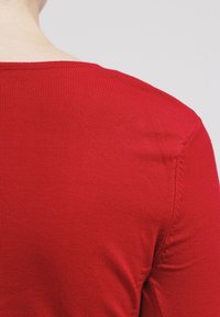 Närbild av en persons övre rygg och axel som bär en röd ribbad stickad tröja mot en neutral ljus bakgrund.