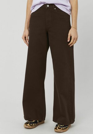 Personne portant un pantalon large marron foncé, un haut lavande clair et des chaussures plates à imprimé léopard, debout devant un fond uni.