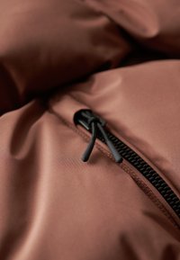 Tissu matelassé marron à la texture lisse avec une fermeture éclair noire et des accents de cordon de serrage, posé sur un fond doux et complémentaire.