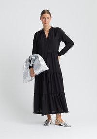 Schwarzes Maxikleid mit langen Ärmeln, gestuftem Design und V-Ausschnitt, kombiniert mit einer metallisch silbernen Clutch und silbernen Ballerinas.