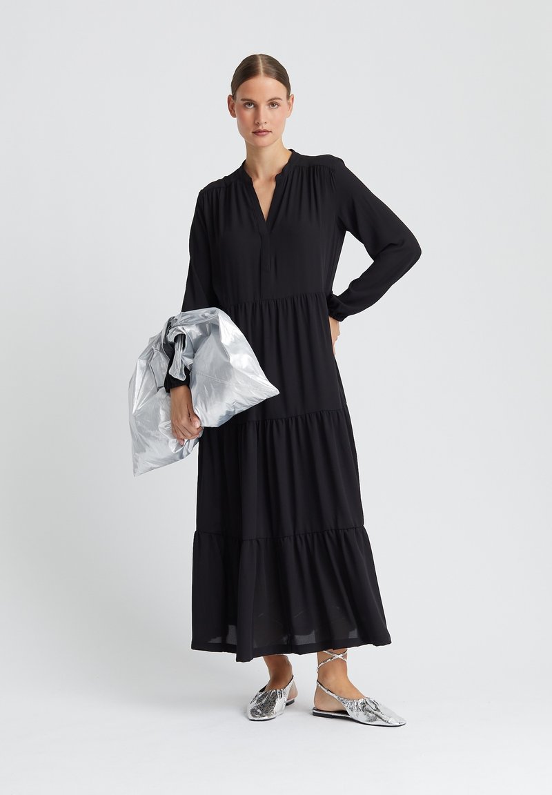 Schwarzes Maxikleid mit langen Ärmeln, gestuftem Design und V-Ausschnitt, kombiniert mit einer metallisch silbernen Clutch und silbernen Ballerinas.