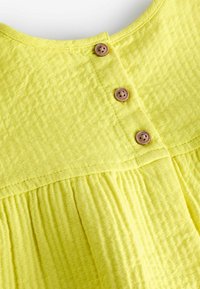 Robe ou haut en tissu texturé jaune avec trois boutons marron à quatre trous le long de la patte de boutonnage avant et une couture froncée en dessous.