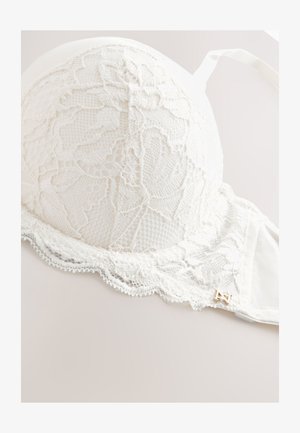 Hvid blonde-bh med blomsterprint, bølgede kanter og en lille guldfarvet dekorativ charme på båndet.