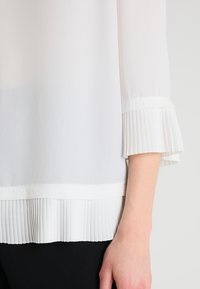 Valkoinen pliseerattu paita, joka on valmistettu pehmeästä, läpikuultavasta kankaasta; siinä on leveät pliseeratut hihat ja pliseerattu alahelma, mikä luo kerroksellisen tekstuurin.