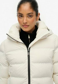 Witte donsjas met een ritssluiting aan de voorkant, voorzien van een hoge kraag en een verborgen capuchon. Draagt over een zwarte, geribbelde coltrui.