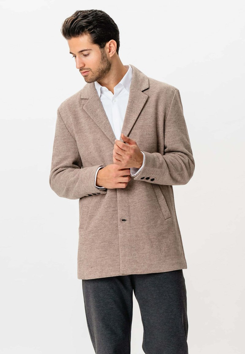 Mand juster beige blazer over hvid skjorte, stående mod en helt hvid baggrund, iført mørkegrå bukser.
