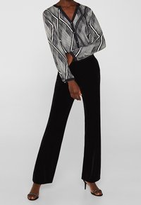 Donna che indossa pantaloni a palazzo in velluto nero, una blusa a fantasia in bianco e nero e sandali neri con tacco e dettagli in strass, in posa su uno sfondo neutro.