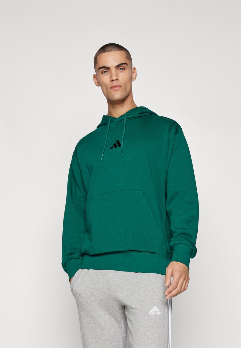 Haut à capuche en coton vert avec une poche avant, capuche à cordon de serrage et logo Adidas noir sur la poitrine. Associé à un pantalon de survêtement gris et une texture lisse.