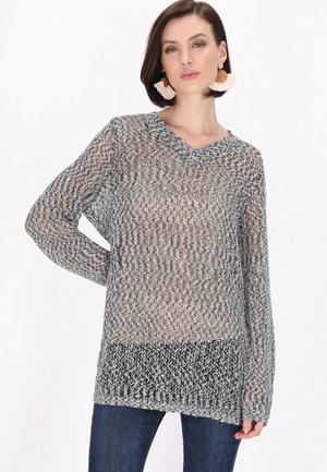 Donna con capelli corti e scuri che indossa grandi orecchini intrecciati e un maglione largo grigio e nero sopra jeans scuri, su sfondo bianco.