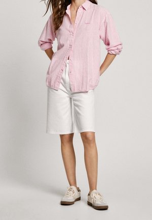 Woman wearing a pink and white striped long-sleeve shirt with sleeves rolled, white knee-length shorts, and white sneakers.