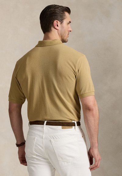 Khaki polo shirt with short sleeves, textured fabric, paired with white jeans and a brown belt. Model facing away, showcasing garment details.
