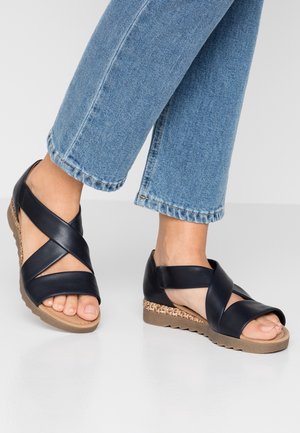 Feet wearing black crisscross leather sandals with tan textured wedge soles paired with light blue denim jeans.