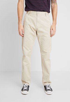 Mężczyzna ubrany w beżowe spodnie o prostych nogawkach, czarno-białe buty sportowe oraz ciemną koszulkę w paski z krótkim rękawem, stojący na jednolitym tle.