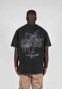 Sort bomulds t-shirt med et stort lilla sommerfugl-grafik på ryggen, med stiliseret tekst og hvide accenter under designet.