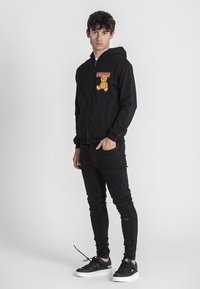 Crna zip-up kapuljača s prednjim džepom, koja sadrži grafiku crtanog medvjeda i natpis "KAWS". U kombinaciji s crnim uskim trapericama i crnim tenisicama.