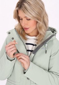 Giacca verde chiaro con cappuccio, colletto interno grigio, chiusura con zip e bottoni a pressione. Sotto, una maglietta a righe. Le mani regolano la giacca.
