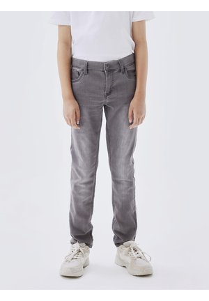 Enfant portant un jean gris délavé, des baskets blanches et une chemise blanche à manches courtes, debout avec les bras détendus le long du corps.