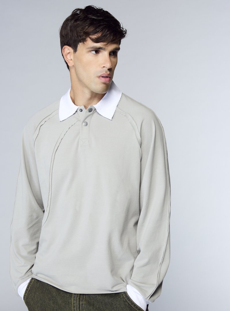 Polo a maniche lunghe grigio chiaro con colletto bianco, caratterizzata da una pattina con bottoni e dettagli di cuciture a contrasto. Tessuto morbido al tatto.