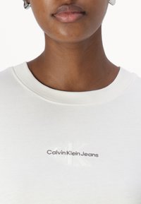 Balta medvilninė marškinėliai su raštuota apykakle, priekyje yra pilku siuvinėjimu užrašytas "Calvin Klein Jeans" logotipas.