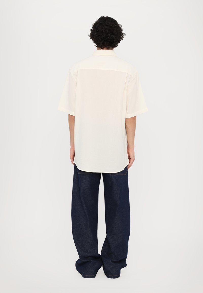 Camisa de manga corta en un color crema claro con un ajuste relajado, combinada con pantalones de mezclilla oscuros de corte holgado, vista desde atrás.